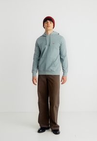 Νεαρός άνδρας που φοράει ανοιχτό μπλε φούτερ με κουκούλα, καφέ παντελόνι, μαύρα παπούτσια και κόκκινο σκουφί, στέκεται μπροστά σε λευκό απλό φόντο.