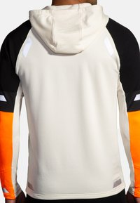 Huvtröja i atletisk stil med krämfärgad kropp, svarta ärmar och klara orange detaljer, med ribbad textur och reflekterande inslag.
