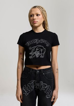 Jeune personne aux cheveux blonds tressés porte un crop top noir et un jean avec des motifs de panthère en contour blanc et le texte "Ed Hardy".
