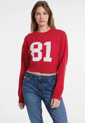Roter, kurzer Pullover mit weißem Design der Zahl "81". Gerippter Rundhalsausschnitt und Bündchen, aus strukturiertem Strickmaterial. Kombiniert mit blauen Jeans.