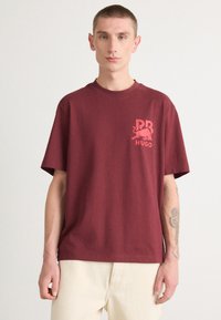 Burgundy cotton t-shirt with short sleeves. Features a light red graphic logo on the left chest. Relaxed fit and crew neck design.