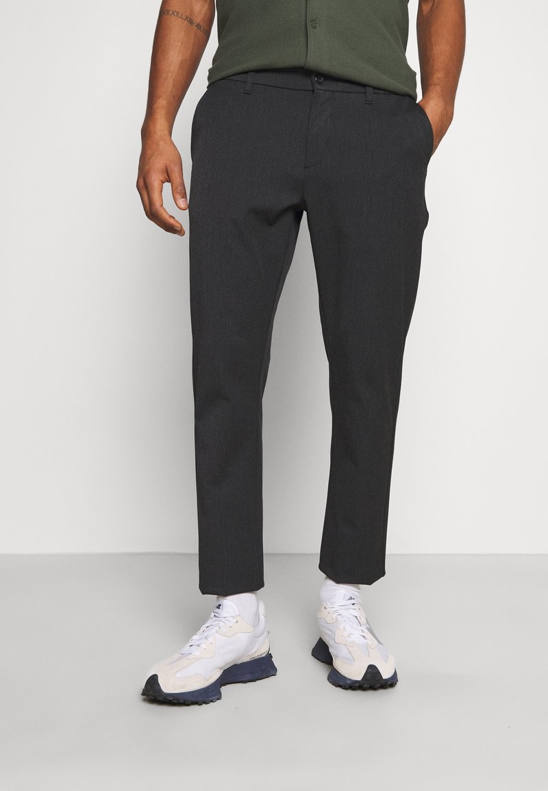 Homme portant un pantalon slim gris foncé, des baskets blanches et une chemise verte, les mains dans les poches, sur un fond uni.