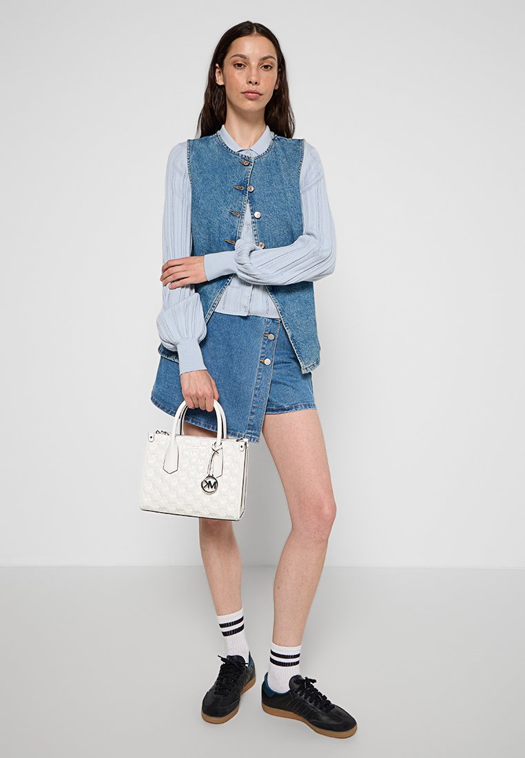 Denimweste über einem hellblauen Langarmoberteil, kombiniert mit einem Denimrock und schwarzen Sneakers. Hält eine weiße Handtasche mit einem Logo-Anhänger.