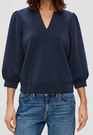 Vrouw die een marineblauwe blouse met V-hals draagt, met geplooide driekwartmouwen en blauwe spijkerbroek, staand met ontspannen armen.