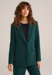 Tyrkysový blazer vyrobený z textilného materiálu, s vykrojeným golierom, dvomi prednými vreckami a dvoma čiernymi gombíkmi. Nosí sa cez čierny top.