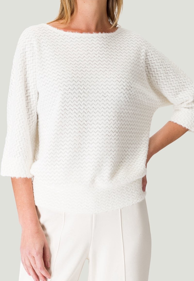 Femme portant un pull en maille blanche texturée en zigzag avec des manches trois-quarts et un pantalon blanc taille haute, main sur la hanche.