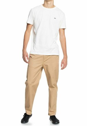 Biała bawełniana koszulka z małym granatowym logo na piersi, zestawiona z khaki spodniami o prostym kroju i subtelnej fakturze. Czarne trampki.