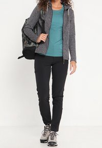 Completo sportivo composto da una giacca grigia con zip e tessuto a trama, una maglietta turchese, pantaloni neri e scarponi da trekking, accompagnati da uno zaino mimetico.