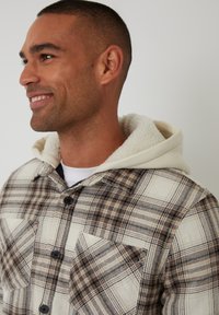 Kurtka w kratę w beżowych i brązowych odcieniach, z miękkim, białym kapturem podszewkowym, z przednimi kieszeniami i czarnymi guzikami.