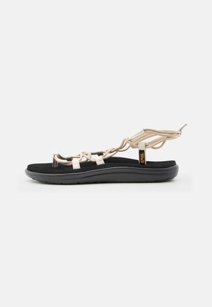 Light beige strappy sandal with black cushioned sole and ankle wrap, featuring a loop toe and Teva brand tag on side.