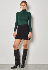 Maglione verde a collo alto, gonna nera a costine e stivali neri in pelle fino al ginocchio con tacco a blocco. Design semplice e aderente.