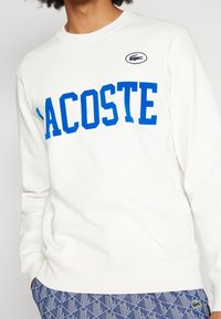 Vit sweatshirt med stor blå "LACOSTE" text över bröstet och en liten svart krokodil-logotyp på övre vänstra sidan.