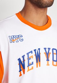 Fehér póló narancssárga és kék kiegészítőkkel. Az elején "NYC" logó található a Nike Swoosh-sal, valamint feltűnő "NEW YORK" felirat grafikus betűtípussal.