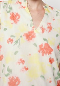 Květinová blůza s bílým podkladem a akvarelovými květinami v červené, žluté a zelené barvě, s výstřihem do V a krátkými rukávy vyrobená z lehkého materiálu.