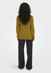 Strickpullover in senfgelb mit einer strukturierten Oberfläche und gerippten Bündchen, kombiniert mit dunklen Jeans im Schlag und weißen Sneakers.