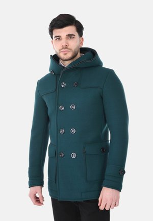 Uomo che indossa un cappotto doppiopetto verde scuro con cappuccio e grandi tasche, in piedi davanti a uno sfondo bianco uniforme.