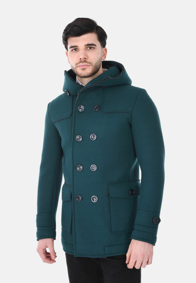 Uomo che indossa un cappotto doppiopetto verde scuro con cappuccio e grandi tasche, in piedi davanti a uno sfondo bianco uniforme.