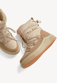Stivali isolati beige con upper in suede e tessuto imbottito, punta tonda e suola in gomma testurizzata. Presentano chiusura con lacci e fodera in pelliccia.