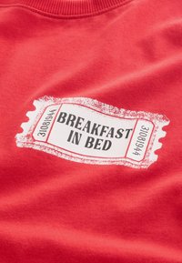 Czerwony materiał z białą grafiką w kształcie biletu, na której znajduje się tekst "ŚNIADANIE DO ŁÓŻKA" oraz liczby po obu stronach.