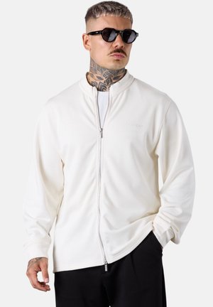 Veste zippée blanche en tissu lisse avec un col montant, accentuée par un logo discret. Le modèle porte des lunettes de soleil noires et un pantalon à coupe décontractée.