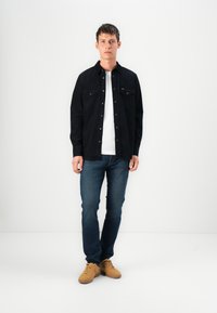 Camisa preta de botão com dois bolsos no peito, usada sobre uma t-shirt branca, combinada com jeans azuis e ténis castanhos sobre um fundo simples.
