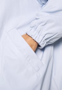 Giacca azzurro chiaro con una texture liscia e lucida. Presenta maniche elastiche e una tasca laterale con orlo cucito.