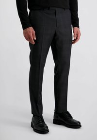 Man in een donkergrijze op maat gemaakte broek en zwarte brogue leren schoenen, staand tegen een effen lichte achtergrond.
