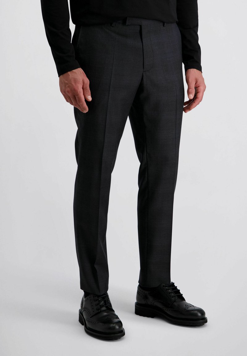 Man in een donkergrijze op maat gemaakte broek en zwarte brogue leren schoenen, staand tegen een effen lichte achtergrond.