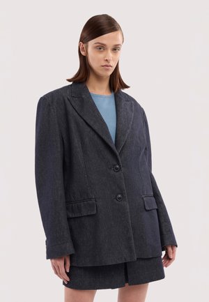 Jonge persoon met steil bruin haar draagt een oversized donkergrijs colbert en bijpassende rok, lichtblauw shirt eronder, staand tegen een neutrale achtergrond.