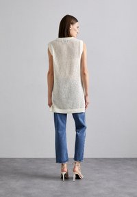 Gilet in maglia beige dalla vestibilità ampia con trama aperta, senza maniche, con spacchi laterali, abbinato a jeans slim blu e décolleté beige con cinturini.