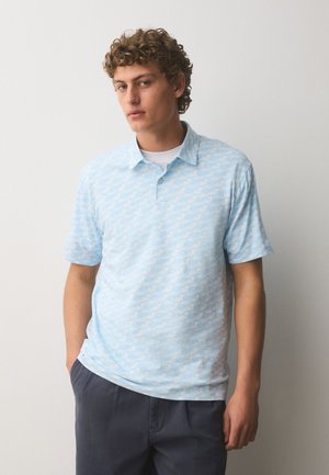 Giovane uomo con capelli ricci che indossa una polo azzurra chiara a fantasia e pantaloni scuri, in piedi davanti a uno sfondo bianco uniforme.