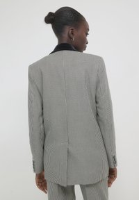 Donna di spalle che indossa un blazer a scacchi bianco e nero e pantaloni abbinati, con i capelli raccolti in un bun e orecchini a cerchio dorati.