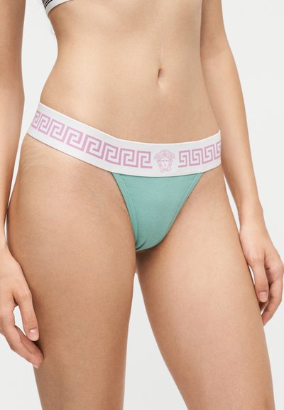 Femme portant un bikini vert avec une ceinture blanche présentant un motif clé grec rose et un dessin de tête de Méduse.