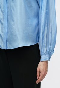 Svetlomodrá, rebrovaná košeľa s dlhým rukávom a zapínaním na gombíky na manžetách, skombinovaná s čiernymi nohavicami. Tkanina pôsobí hladko a mierne leskle.