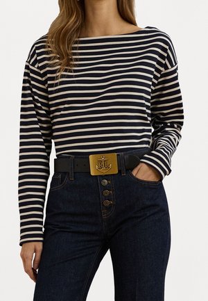 Femme portant un chemisier à manches longues rayé bleu marine et blanc, un jean taille haute foncé avec braguette à boutons, et une ceinture noire avec une boucle en forme d'ancre dorée.