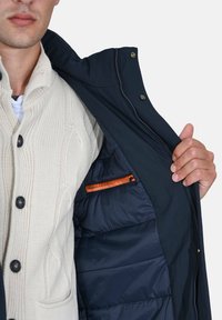 Cappotto blu navy con fodera interna strutturata, dotato di tasca con cerniera arancione. Tessuto esterno liscio; bottoni visibili e dettaglio del colletto.