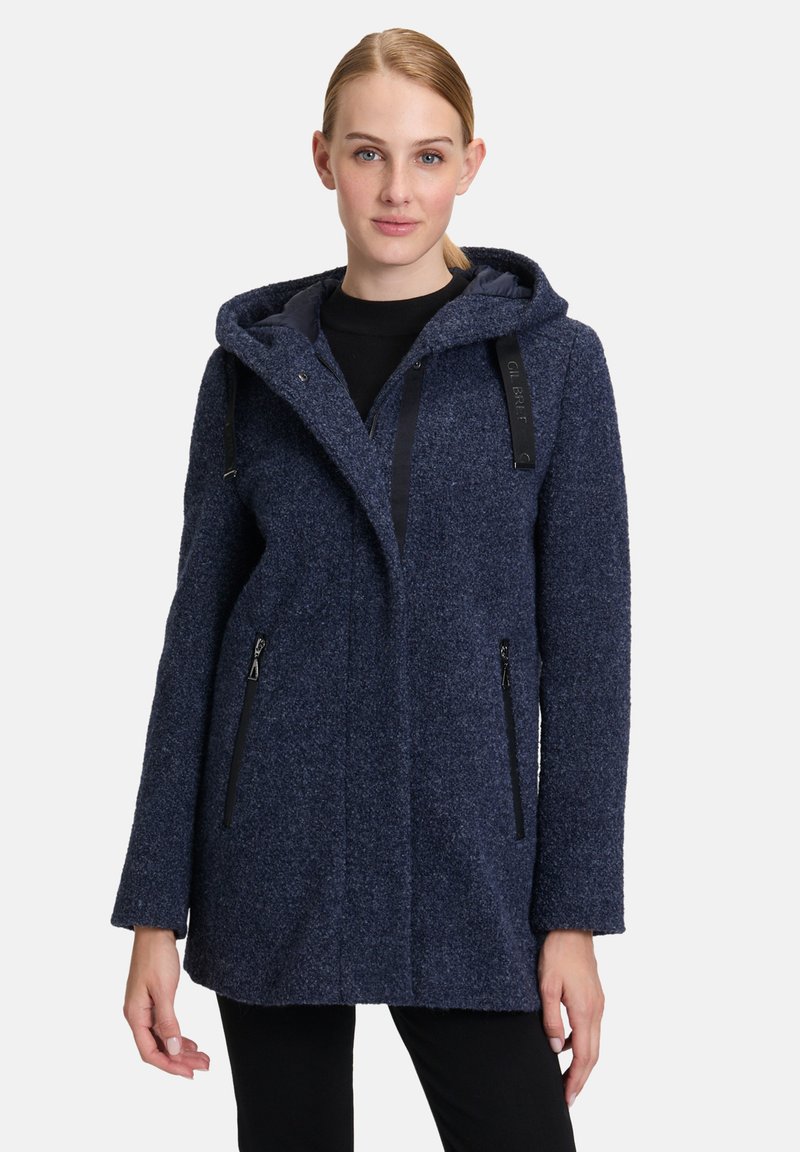 Granatowy, teksturowany płaszcz z kapturem. Posiada zapinane na zamek kieszenie z przodu oraz regulowane paski. Luźny krój z prostym sylwetką.