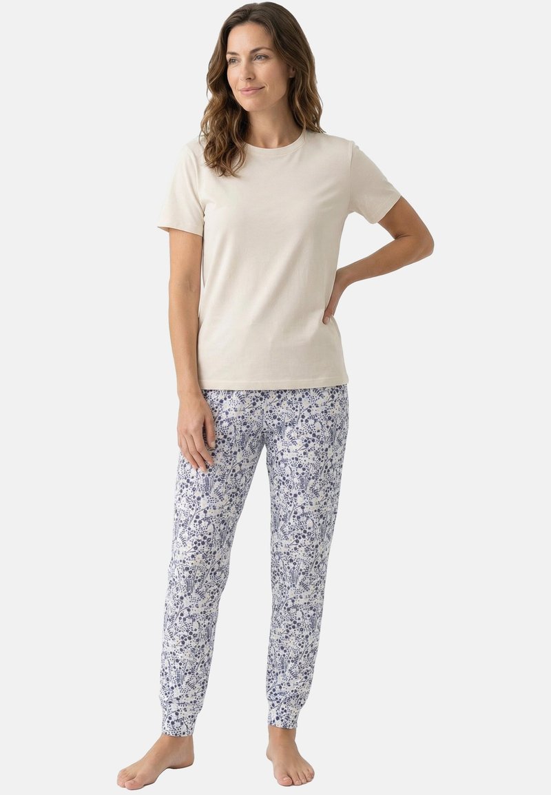 Femme debout pieds nus portant un t-shirt beige à manches courtes et un pantalon blanc à motif floral sur un fond uni.
