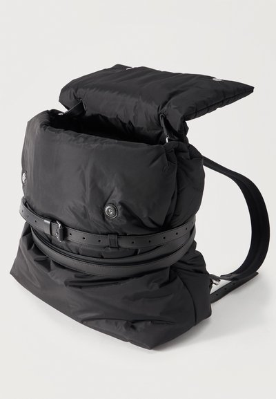 Μαύρο μαξιλαρωμένο σακίδιο από νάιλον με ανοιχτό καπάκι στην κορυφή, κουμπιά με πατιλέτα μπροστά και δύο λεπτά ρυθμιζόμενα δερμάτινα λουριά γύρω από το σώμα.