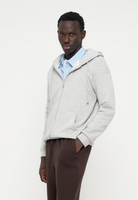Grau zip-up Kapuzenpullover mit Kordelzügen, der über einem hellblau gestreiften Shirt getragen wird und mit braunen Jogginghosen kombiniert ist.