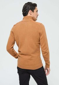 Pull à col montant en tricot côtelé de couleur marron chaud avec des accents aux épaules. Design ajusté avec des manches longues et un col légèrement montant.