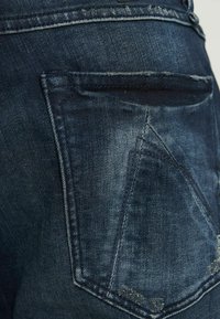 Temno modri jeans iz denima z zadnjim žepom z značilnim trikotnim dizajnom in rahlim bledenjem. Šivi so vidni ob robovih.