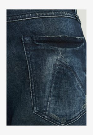 Tumed sinise denimist teksad, millel on seljatasku eristuva kolmnurga kujundusega ja õrn fade. Õmblused on nähtavad servadel.