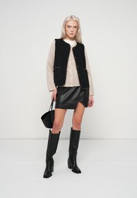 Μαύρο υφασμάτινο γιλέκο πάνω από ένα μπεζ πουλόβερ, σε συνδυασμό με μια μαύρη δερμάτινη μίνι φούστα και μαύρες ψηλές μπότες με τακούνι. Μικρή μαύρη τσάντα χειρός.