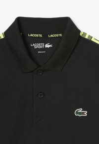 Polo nero in tessuto testurizzato, con tre bottoni e logo di coccodrillo verde. Accenti giallo neon sul colletto e sulla spalla.