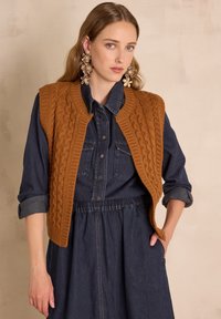 Gilet en tricot marron avec un motif de torsades sur une robe chemise en denim foncé, dotée d'une taille froncée et de deux poches poitrine.