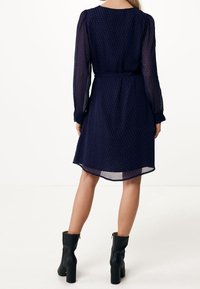 Vestido azul marino con un patrón geométrico, mangas largas y transparentes, cintura entallada y falda ligeramente acampanada, acompañado de botas negras hasta el tobillo.