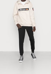 Ljusbeige lättviktsjacka med halvlång dragkedja, stor framficka och svart logotyp. Matchad med svarta joggingbyxor och högstövlar.