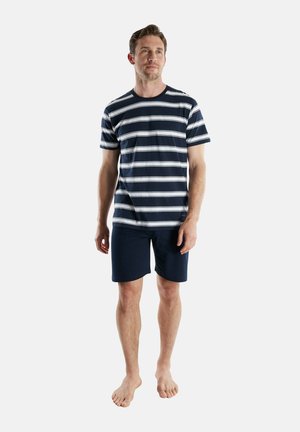 Mann steht barfuß, trägt ein navyblau-weiß gestreiftes T-Shirt und navyblaue Shorts vor einem einfarbigen Hintergrund.