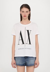 White cotton t-shirt with short sleeves, featuring a large black "AX" logo and "ARMANI EXCHANGE" text below. Styled with denim shorts.
